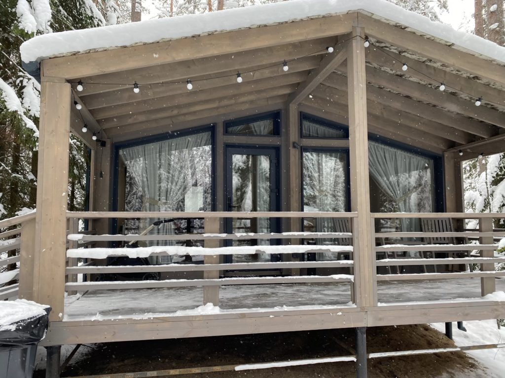 Деревянный домик, вдохновленный домом из клееного бруса, с большими стеклянными окнами и крытым крыльцом стоит в заснеженном лесу. С крыши свисают гирлянды, снег покрывает перила и землю в окружении высоких деревьев.